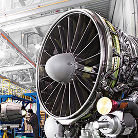 A huge aircraft engine sits inside a bright factory while engineers inspect other aircraft components in the background
