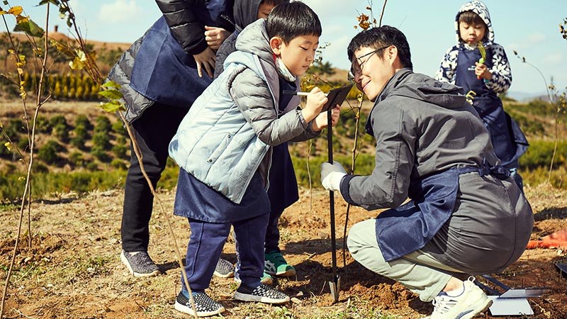 On a sunny day, a young boy writes a message for Hanwha Solar Forest visitors on a card while surrounded by friends