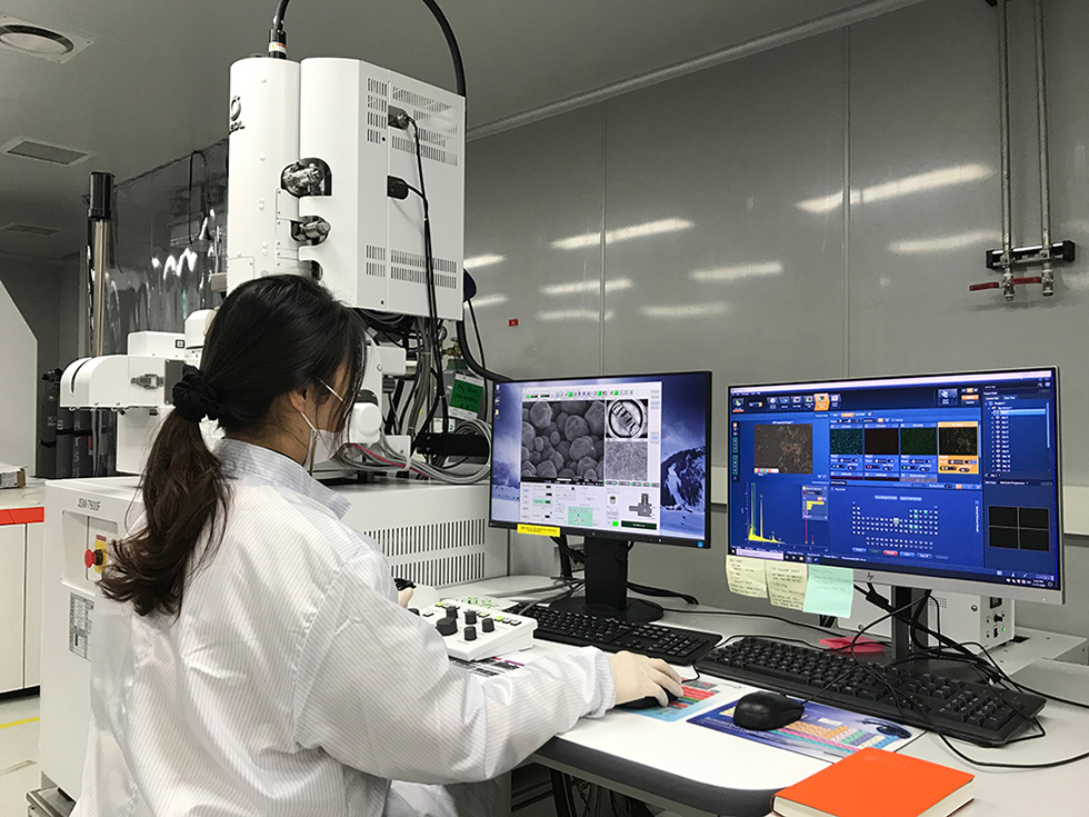 A single researcher in a white lab coat looking at two desktop monitors with research data on-screen in a lab surrounded by machinery