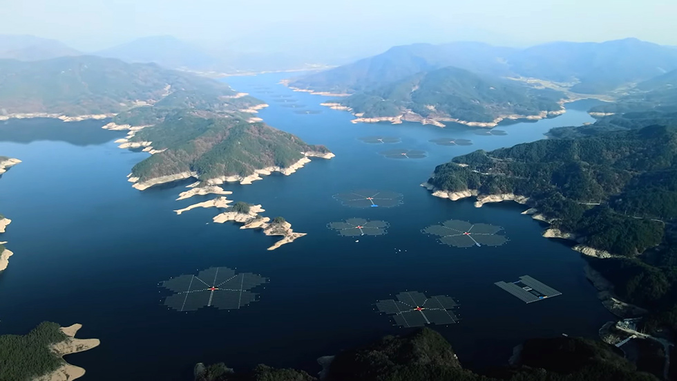An aerial photo of several flower-shaped solar PV groupings floating on a dark blue lake surrounded by rolling green hills