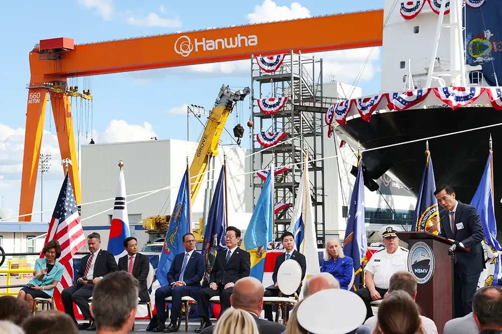 Several leaders sit in front of Hanwha Philly Shipyard in front of an audience during a christening ceremony.