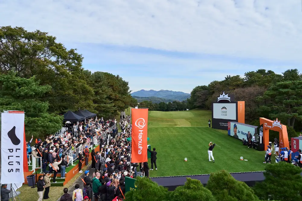 A golfer tees off in front of LIFEPLUS and Hanwha banners as spectators watch.