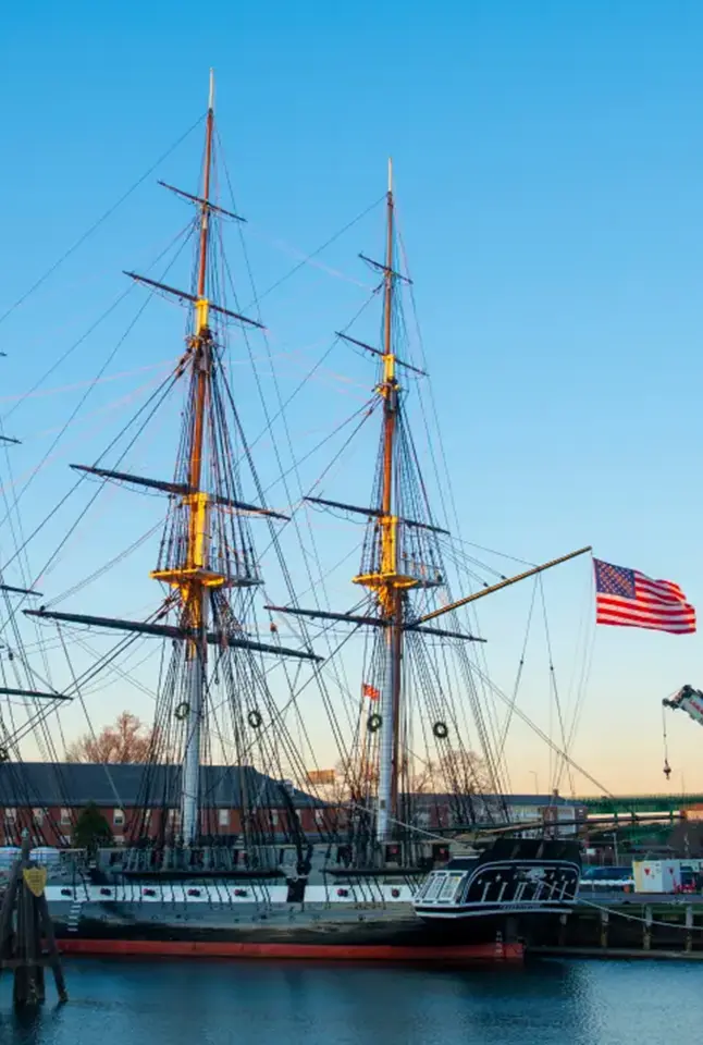 Ship with tall masts docked in harbor as the American flag waves at sunset.