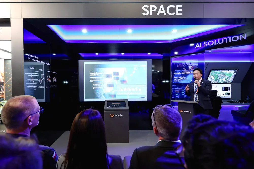 A man speaks to an audience at a large Hanwha booth showcasing exhibits on space and AI solutions.